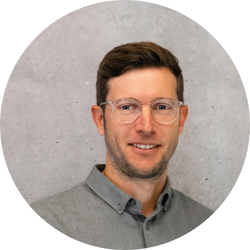 Porträt eines Mannes mit Brille, lächelnd vor einem grauen Hintergrund. Er trägt ein helles Hemd.