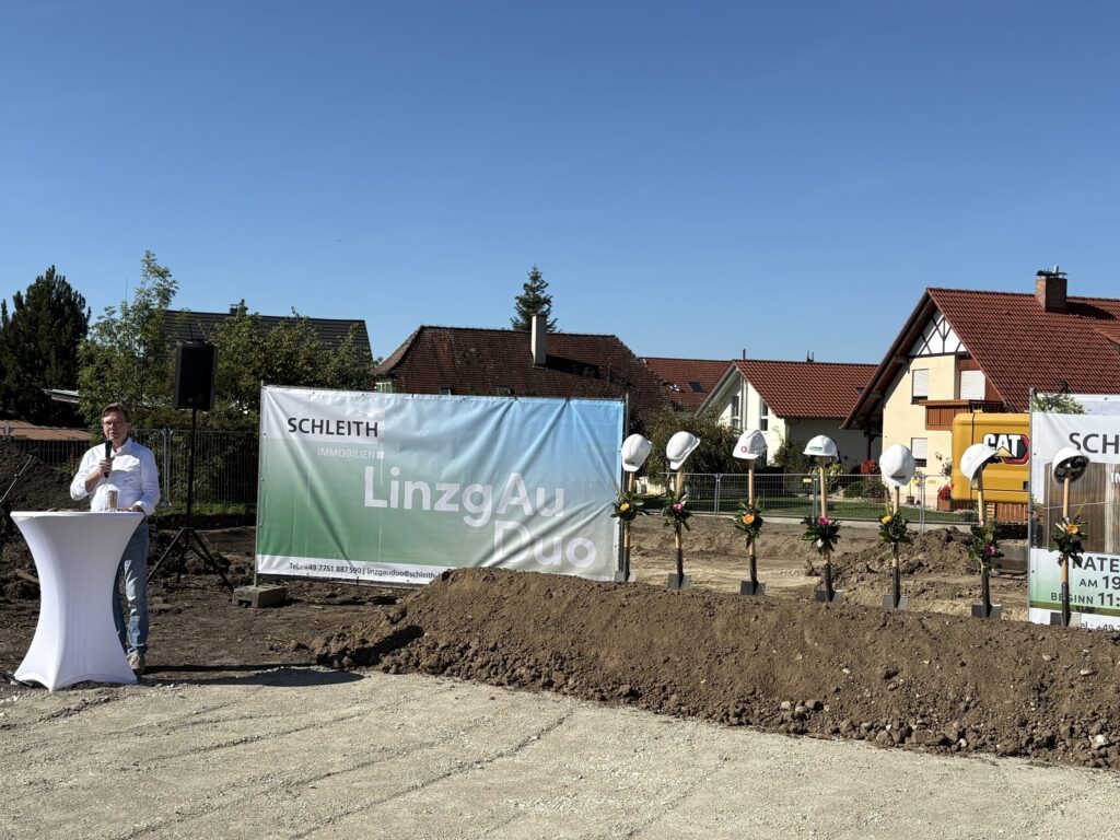 Ansprache beim Spatenstich des LinzgAuDuo in Frickingen vor dem Banner von SCHLEITH Immobilien, daneben die aufgereihten Spaten mit Blumenschmuck.