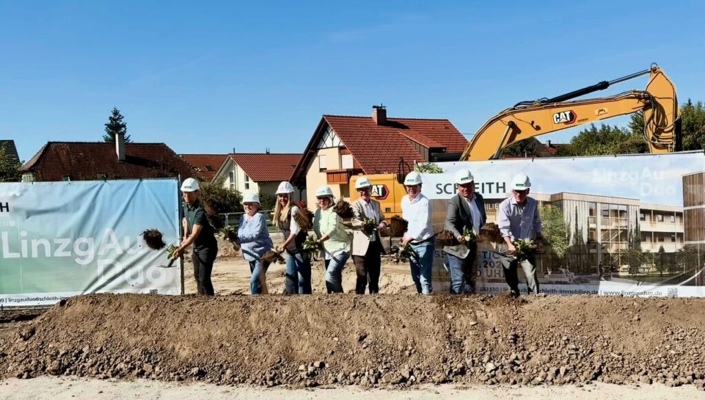 Spatenstich für das Wohnprojekt LinzgAuDuo in Frickingen: Beteiligte mit Bauhelmen und Spaten vor dem Bauzaun, im Hintergrund ein Bagger.